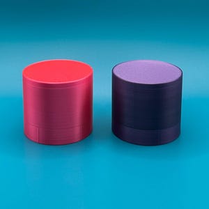 Op de afbeelding: Twee cilindervormige houders met platte bovenkanten. De houder aan de linkerkant is fel magentarood, terwijl de houder aan de rechterkant diep donkerpaars is. Beide objecten hebben een gladde, effen gekleurde buitenkant en een bijpassend rond deksel.