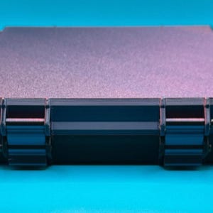 May include: A rectangular, dark-colored storage case with a textured, matte-finish lid and two prominent, sturdy hinges on the front edge. The case features a sleek, low-profile design with clean lines and a solid, opaque construction.