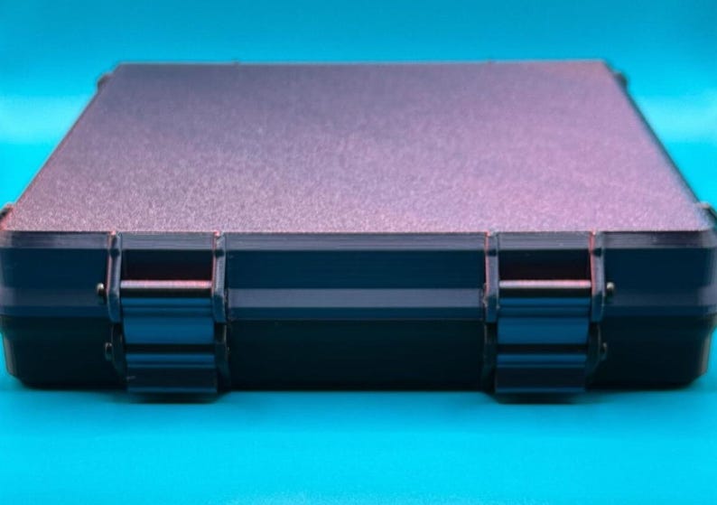 May include: A rectangular, dark-colored storage case with a textured, matte-finish lid and two prominent, sturdy hinges on the front edge. The case features a sleek, low-profile design with clean lines and a solid, opaque construction.