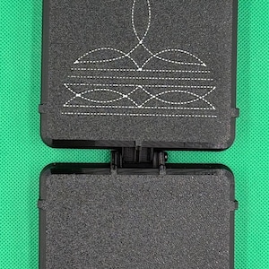May include: A black square quilting template with a textured surface and a hinged design. The top section features a white stitched-style pattern of a decorative crown or floral motif, while the bottom section remains plain and solid black.