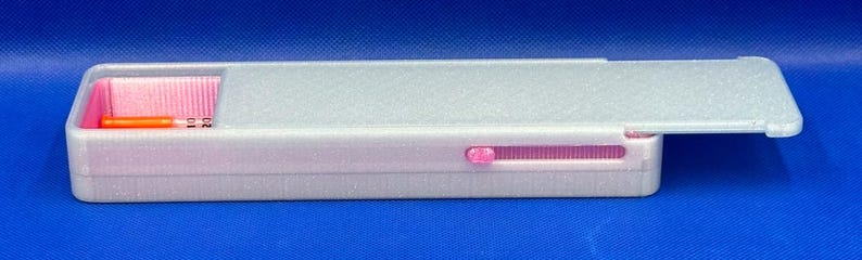 May include: A rectangular white pencil case with a sliding lid and a pink interior. The case features a small pink sliding mechanism on the side and contains a single orange pencil visible in the open compartment on the left side.