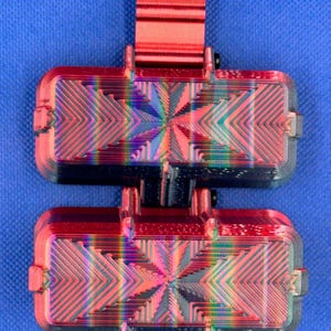 May include: A dual-component rectangular device featuring a metallic red top clip and two stacked, horizontally oriented rectangular blocks. Each block displays a vibrant, iridescent rainbow-colored geometric pattern with sharp, angular lines.