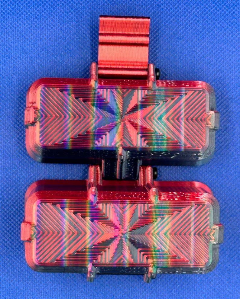 May include: A dual-component rectangular device featuring a metallic red top clip and two stacked, horizontally oriented rectangular blocks. Each block displays a vibrant, iridescent rainbow-colored geometric pattern with sharp, angular lines.