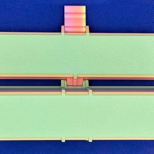May include: A rectangular, two-part hinged container featuring a light green textured surface on both lids. The frame is constructed with a gradient of pink, orange, and yellow colors. The object is symmetrical with a central hinge mechanism.