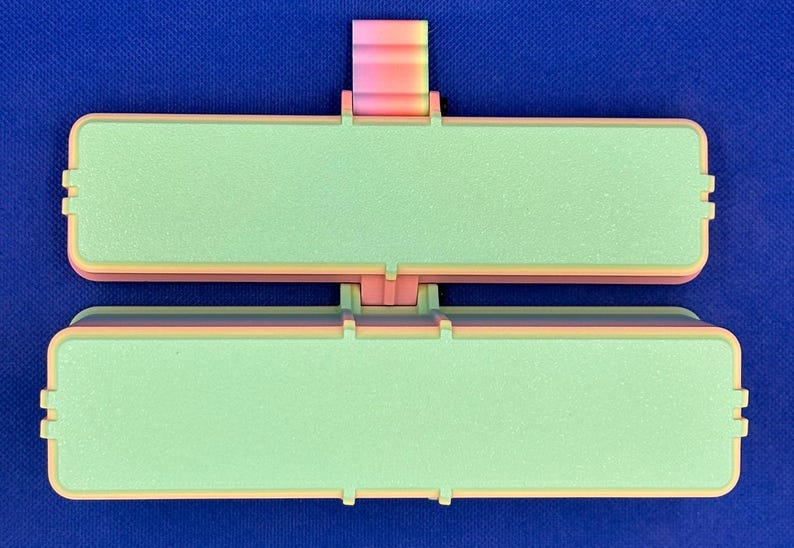 May include: A rectangular, two-part hinged container featuring a light green textured surface on both lids. The frame is constructed with a gradient of pink, orange, and yellow colors. The object is symmetrical with a central hinge mechanism.