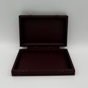 May include: An open, rectangular, burgundy-colored box with a hinged lid. The box is made of a solid material and has a simple, classic design. The lid is open, revealing the interior of the box.