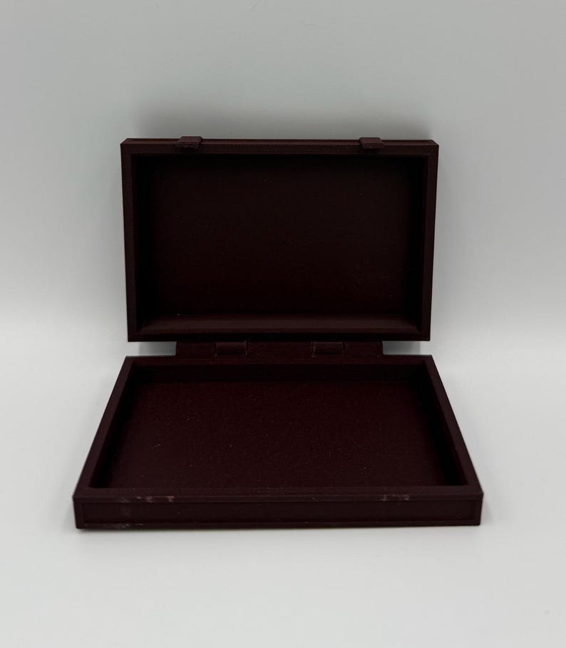 May include: An open, rectangular, burgundy-colored box with a hinged lid. The box is made of a solid material and has a simple, classic design. The lid is open, revealing the interior of the box.