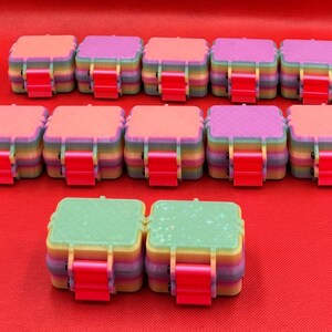 May include: Twelve square storage containers with rainbow-colored sides and bright pink latches. The lids feature various textures, including solid colors, cross-hatch patterns, and a light green surface with a subtle star-shaped glitter effect.