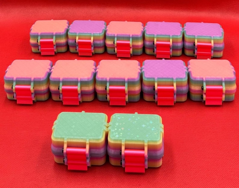 May include: Twelve square storage containers with rainbow-colored sides and bright pink latches. The lids feature various textures, including solid colors, cross-hatch patterns, and a light green surface with a subtle star-shaped glitter effect.