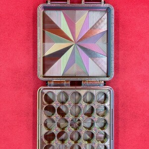 May include: A rectangular, hinged storage case with a multi-colored, iridescent finish. The top interior features a geometric starburst pattern, while the bottom interior contains a grid of twenty-five circular compartments.