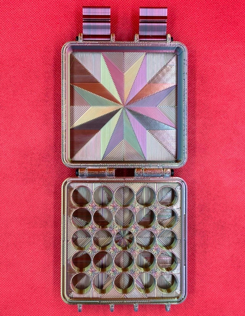 May include: A rectangular, hinged storage case with a multi-colored, iridescent finish. The top interior features a geometric starburst pattern, while the bottom interior contains a grid of twenty-five circular compartments.