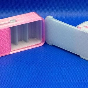May include: Two rectangular sliding cases with internal dividers. The left case is pink with a textured lid and gray interior. The right case is white with a smooth gray lid and a pink interior. Both feature a small sliding mechanism on top.