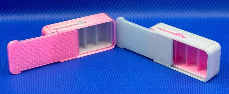 May include: Two rectangular sliding cases with internal dividers. The left case is pink with a textured lid and gray interior. The right case is white with a smooth gray lid and a pink interior. Both feature a small sliding mechanism on top.