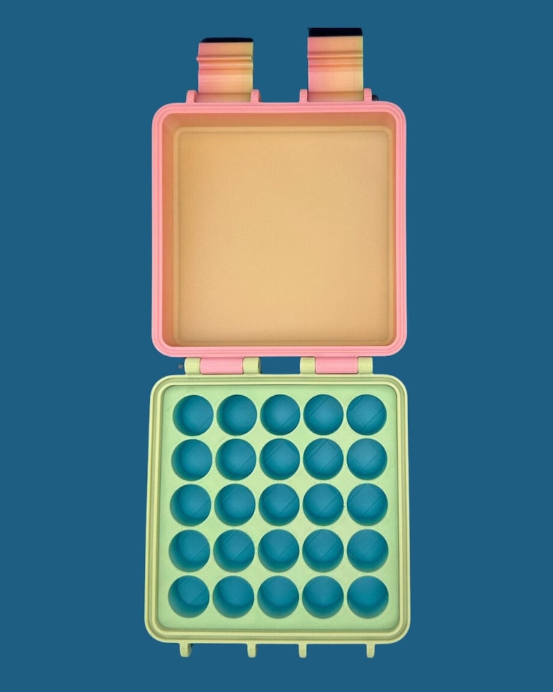 May include: A square, open storage case featuring a light pink lid and a light green base. The interior of the base contains a grid of twenty-five circular indentations in a bright blue color. Two hinges are visible at the top edge.