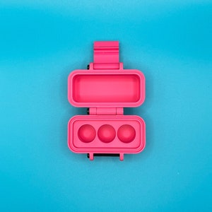 May include: A pink plastic container with three round compartments, open to reveal the empty compartments.