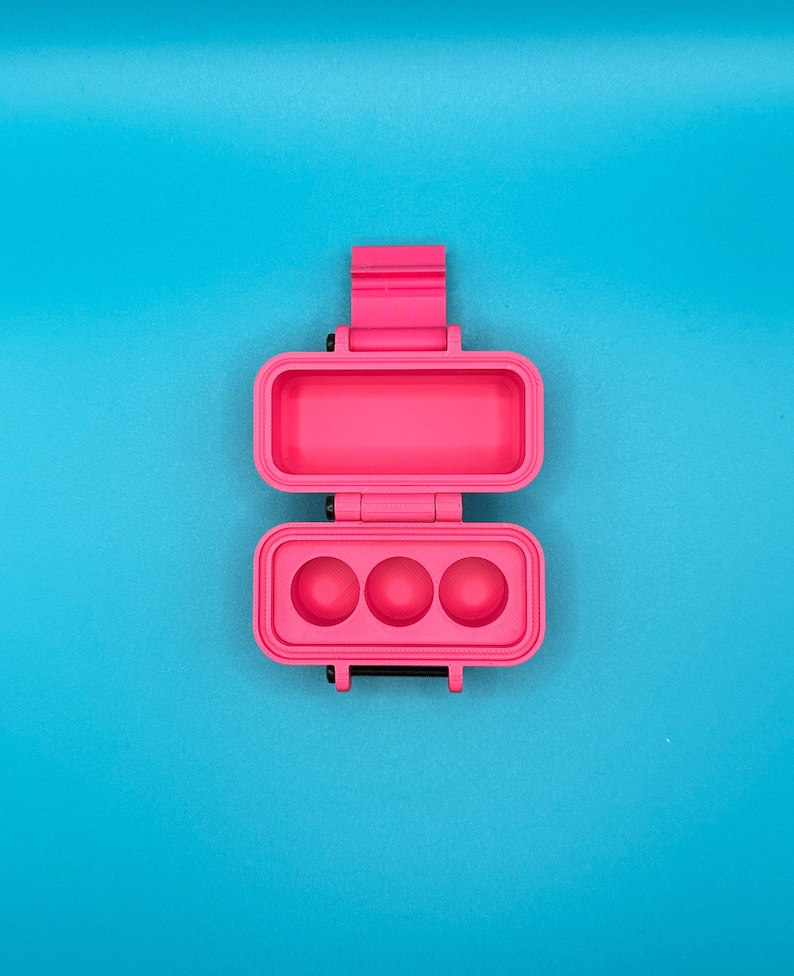 Puede incluir: Un estuche de almacenamiento rectangular de color rosa brillante con bisagras, hecho de un material s&oacute;lido. El estuche est&aacute; abierto, revelando un compartimento superior hueco y una secci&oacute;n inferior con tres hendiduras circulares.