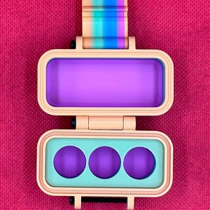 Puede incluir: Una caja rectangular de pl&aacute;stico beige con bisagras y un cierre de rayas arco&iacute;ris. La secci&oacute;n superior presenta un interior rectangular de color morado intenso, mientras que la secci&oacute;n inferior contiene un inserto verde claro con tres aberturas circulares moradas.