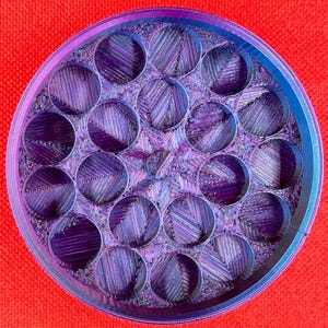 May include: A circular container with a multi-compartment interior featuring a grid of nineteen cylindrical openings. The object displays a gradient of iridescent purple and blue tones with a textured, layered finish throughout the structure.