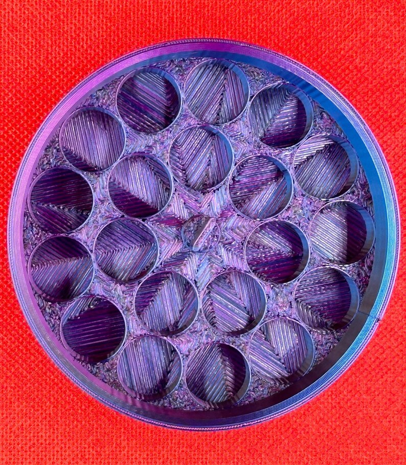May include: A circular container with a multi-compartment interior featuring a grid of nineteen cylindrical openings. The object displays a gradient of iridescent purple and blue tones with a textured, layered finish throughout the structure.