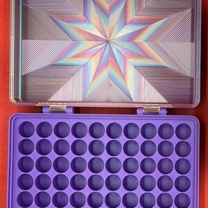 May include: A rectangular storage case with a purple interior featuring a grid of sixty circular compartments. The lid interior displays a multi-colored, iridescent starburst pattern with geometric lines, and the hinges feature a plaid fabric.
