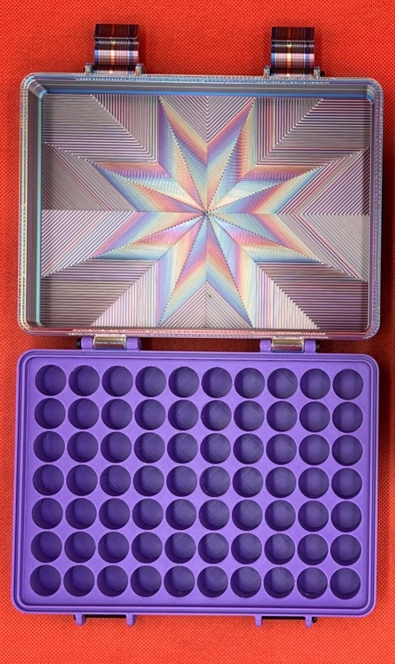 May include: A rectangular storage case with a purple interior featuring a grid of sixty circular compartments. The lid interior displays a multi-colored, iridescent starburst pattern with geometric lines, and the hinges feature a plaid fabric.
