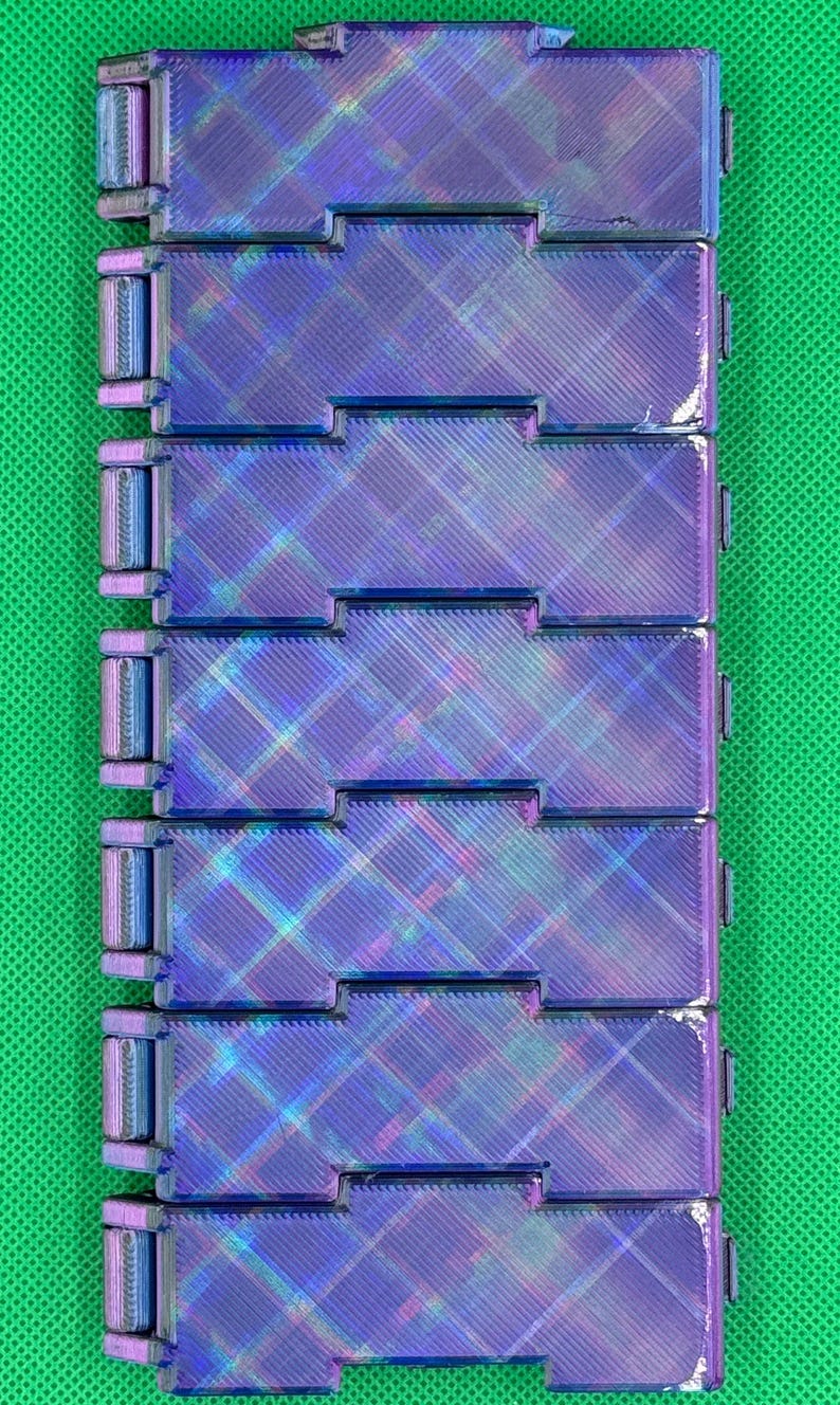 May include: A rectangular, articulated fidget toy composed of seven interconnected, iridescent purple and blue plastic segments. Each segment features a textured, diagonal cross-hatch pattern on its surface, creating a metallic, shifting light effect across the entire linked structure.
