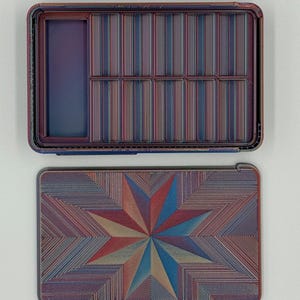 May include: An open, rectangular box with a lid. The box and lid are decorated with a repeating pattern of thin lines in shades of red, blue, and tan. The lid features a starburst design.