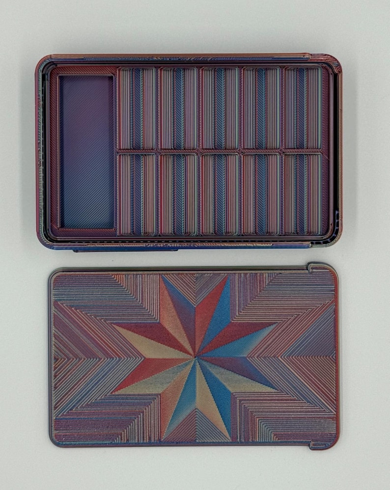 May include: An open, rectangular box with a lid. The box and lid are decorated with a repeating pattern of thin lines in shades of red, blue, and tan. The lid features a starburst design.