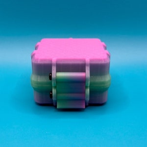 May include: A square, multi-colored storage box featuring a gradient transition from pink on the top lid to a light purple and green base. The container includes a central latch mechanism with two small black pins on the front side.