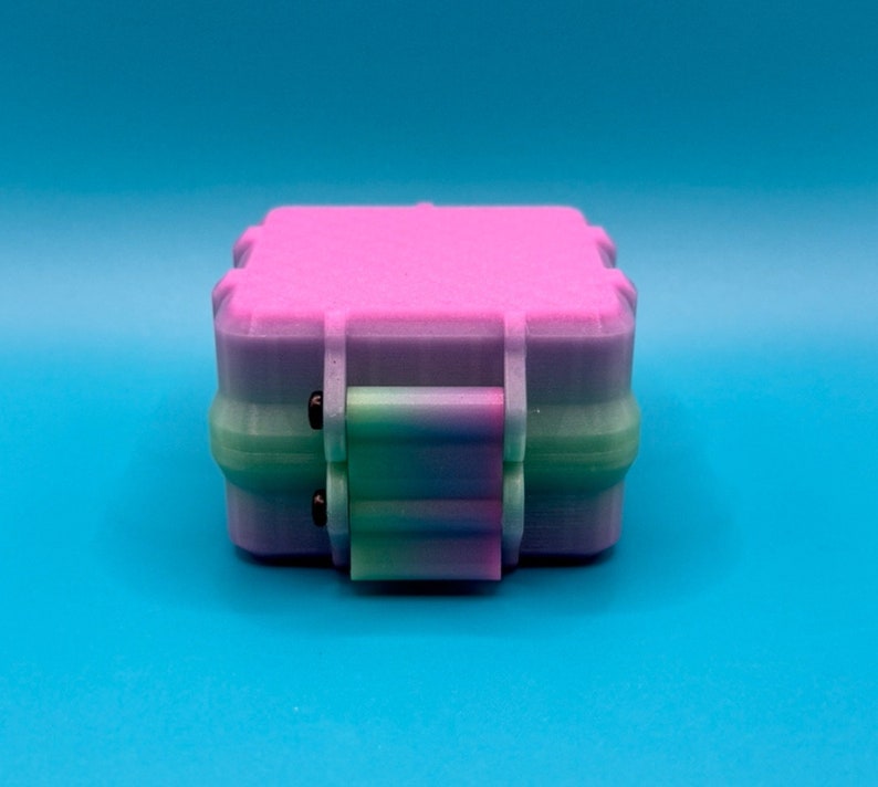 May include: A square, multi-colored storage box featuring a gradient transition from pink on the top lid to a light purple and green base. The container includes a central latch mechanism with two small black pins on the front side.