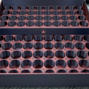 May include: A two-part, open storage tray with numerous circular compartments. The tray is primarily dark purple with a reddish-brown interior, and is designed to hold small cylindrical objects. The top part of the tray is open.