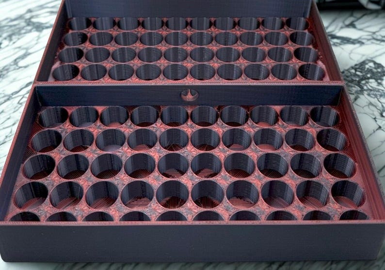 May include: A two-part, open storage tray with numerous circular compartments. The tray is primarily dark purple with a reddish-brown interior, and is designed to hold small cylindrical objects. The top part of the tray is open.