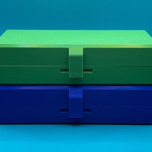 May include: A stack of two rectangular storage boxes with hinged lids and central latch closures. The top box is bright green with a textured lid surface, while the bottom box is solid blue. Both containers feature a smooth, matte finish.