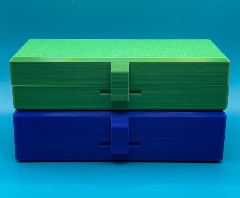 May include: A stack of two rectangular storage boxes with hinged lids and central latch closures. The top box is bright green with a textured lid surface, while the bottom box is solid blue. Both containers feature a smooth, matte finish.