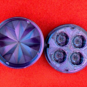 Op de afbeelding: Een ronde, tweedelige kruidenmolen met een iriserende paarse en blauwe afwerking. Het bovendeel heeft een geometrisch, stervormig patroon van ribbels, terwijl het onderste deel vier ronde kamers bevat met zichtbare metalen tanden binnenin.