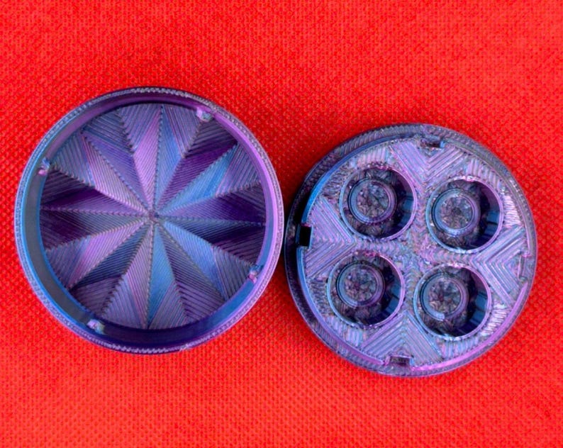 Op de afbeelding: Een ronde, tweedelige kruidenmolen met een iriserende paarse en blauwe afwerking. Het bovendeel heeft een geometrisch, stervormig patroon van ribbels, terwijl het onderste deel vier ronde kamers bevat met zichtbare metalen tanden binnenin.