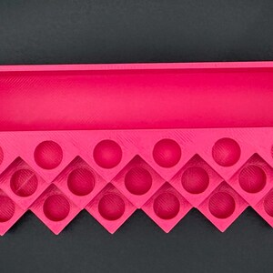 May include: A bright pink 3D-printed organizer tray featuring a long rectangular storage compartment at the top and a series of circular cutouts arranged in a zigzag pattern along the bottom section for holding small cylindrical items.
