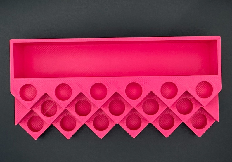 May include: A bright pink 3D-printed organizer tray featuring a long rectangular storage compartment at the top and a series of circular cutouts arranged in a zigzag pattern along the bottom section for holding small cylindrical items.