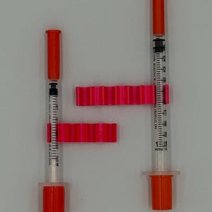 May include: Two insulin syringes with orange caps and plungers, alongside two bright pink, ridged syringe holders. The syringes are marked with measurements in ml/cc. The image is set against a plain white background.
