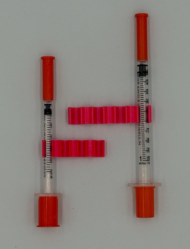 May include: Two insulin syringes with orange caps and plungers, alongside two bright pink, ridged syringe holders. The syringes are marked with measurements in ml/cc. The image is set against a plain white background.
