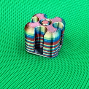 May include: A 3D printed cube-shaped object with rounded corners and a central circular indentation. The surface features horizontal layers of iridescent, multi-colored filament in shades of blue, red, yellow, and green throughout the structure.