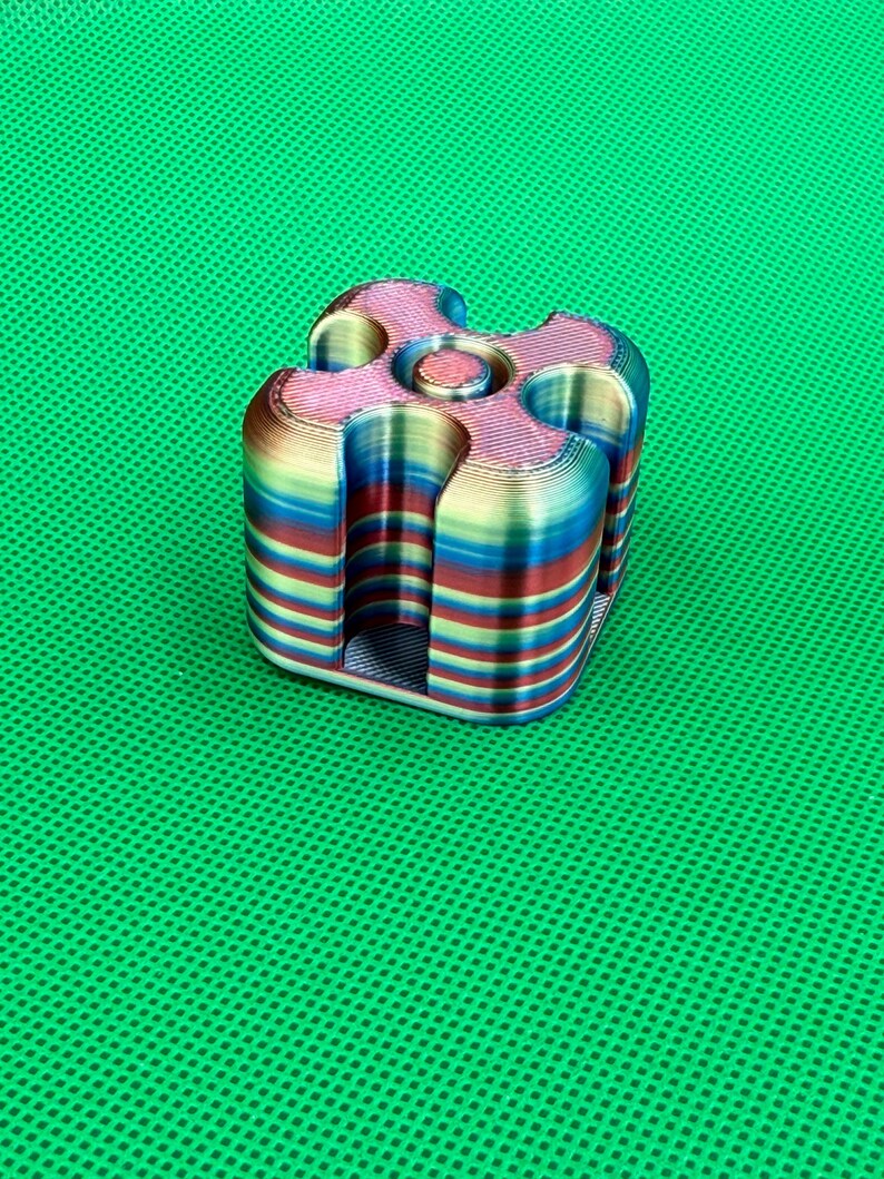 May include: A 3D printed cube-shaped object with rounded corners and a central circular indentation. The surface features horizontal layers of iridescent, multi-colored filament in shades of blue, red, yellow, and green throughout the structure.