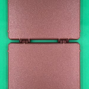 K&ouml;nnte beinhalten: Ein quadratisches, braunes Kunststoffgeh&auml;use mit Scharnier und strukturierter, matter Oberfl&auml;che. Das Objekt besteht aus zwei identischen flachen Platten, die durch einen zentralen Scharniermechanismus verbunden sind, mit zwei kleinen Verschlusslaschen.