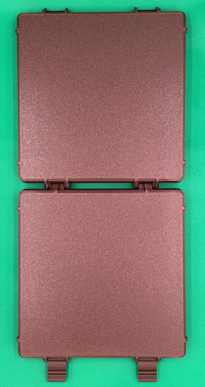 K&ouml;nnte beinhalten: Ein quadratisches, braunes Kunststoffgeh&auml;use mit Scharnier und strukturierter, matter Oberfl&auml;che. Das Objekt besteht aus zwei identischen flachen Platten, die durch einen zentralen Scharniermechanismus verbunden sind, mit zwei kleinen Verschlusslaschen.
