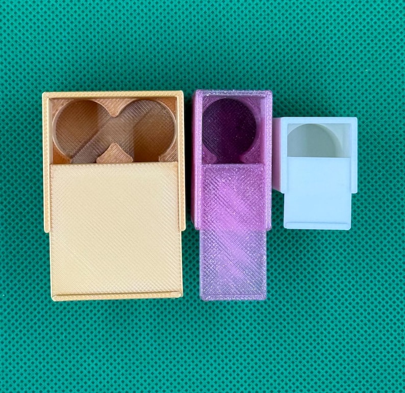 May include: Three 3D printed plastic containers of varying sizes and colors. The largest is a light tan rectangular box with a double circular cutout. The middle is a translucent purple rectangular box. The smallest is a solid white box.