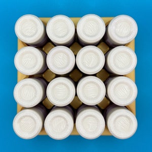 May include: A square yellow cardboard tray holds sixteen cylindrical containers arranged in a four-by-four grid. Each container features a white circular lid with a textured, lined surface pattern, viewed from directly above.