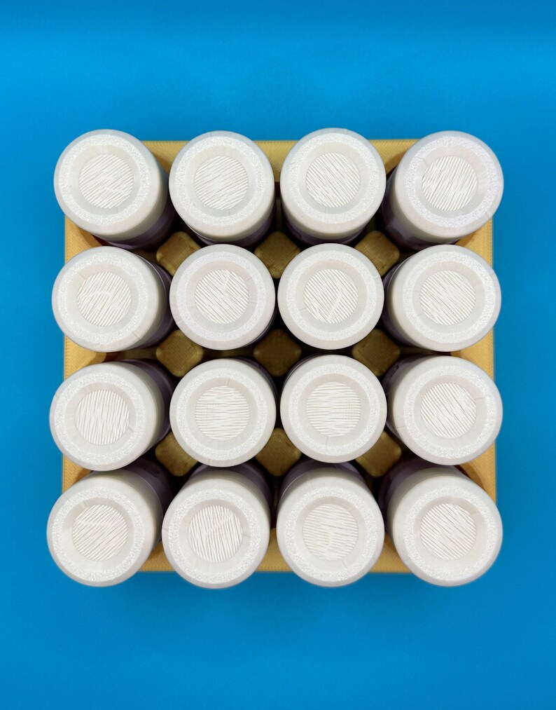 May include: A square yellow cardboard tray holds sixteen cylindrical containers arranged in a four-by-four grid. Each container features a white circular lid with a textured, lined surface pattern, viewed from directly above.