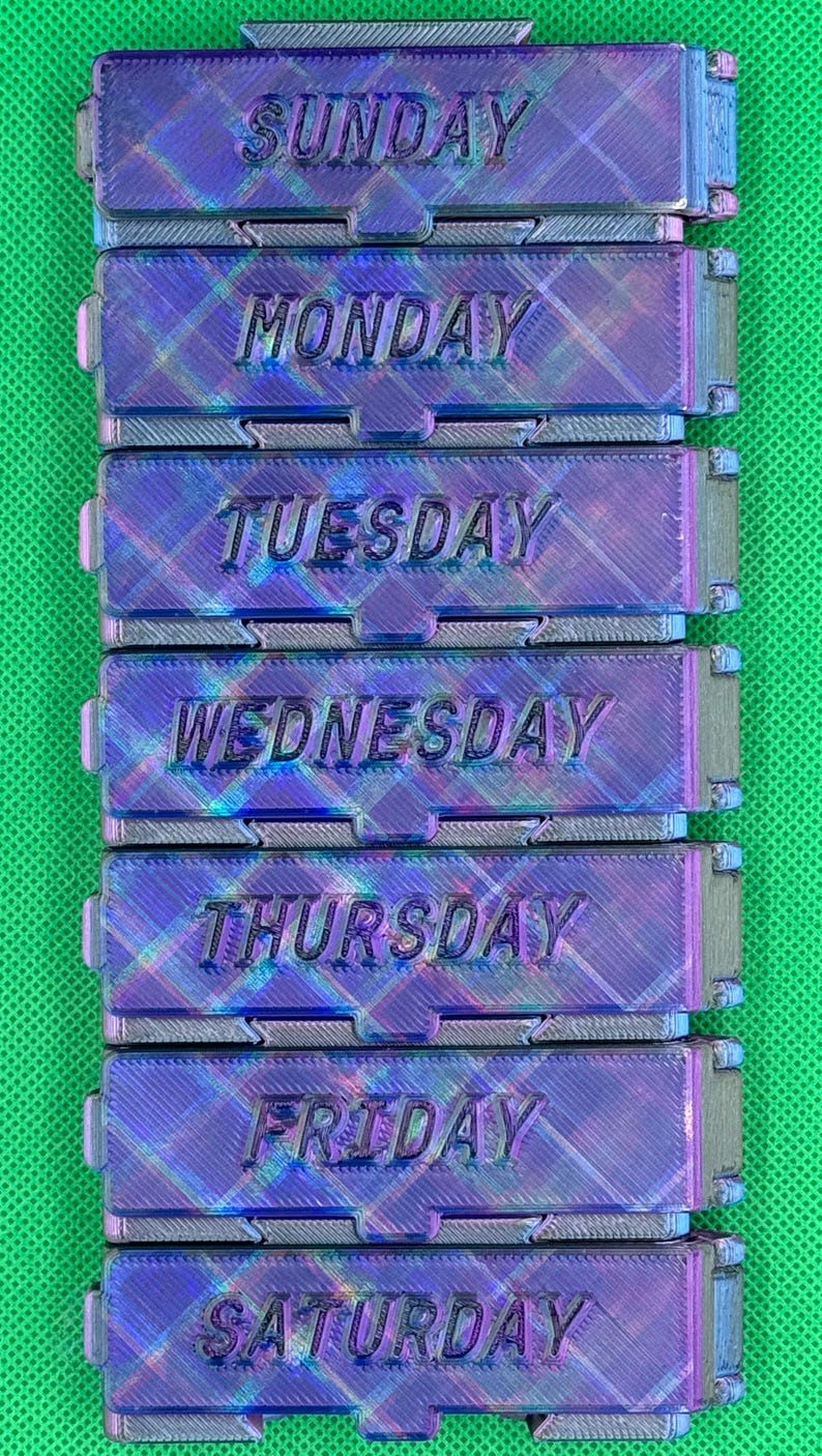 May include: A set of seven rectangular, iridescent purple and blue plastic containers stacked vertically. Each container features a hinged lid embossed with the days of the week: Sunday, Monday, Tuesday, Wednesday, Thursday, Friday, and Saturday.
