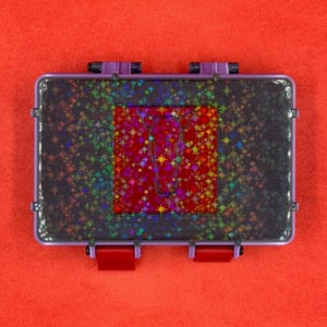 May include: A rectangular, clear plastic case with a purple frame and red clasps. The case has a dark, glittery surface with a central red square adorned with holographic stars and a cursive design. The background is a solid orange.