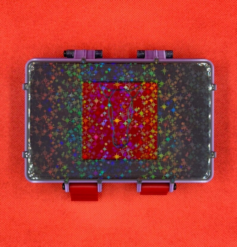 May include: A rectangular, clear plastic case with a purple frame and red clasps. The case has a dark, glittery surface with a central red square adorned with holographic stars and a cursive design. The background is a solid orange.